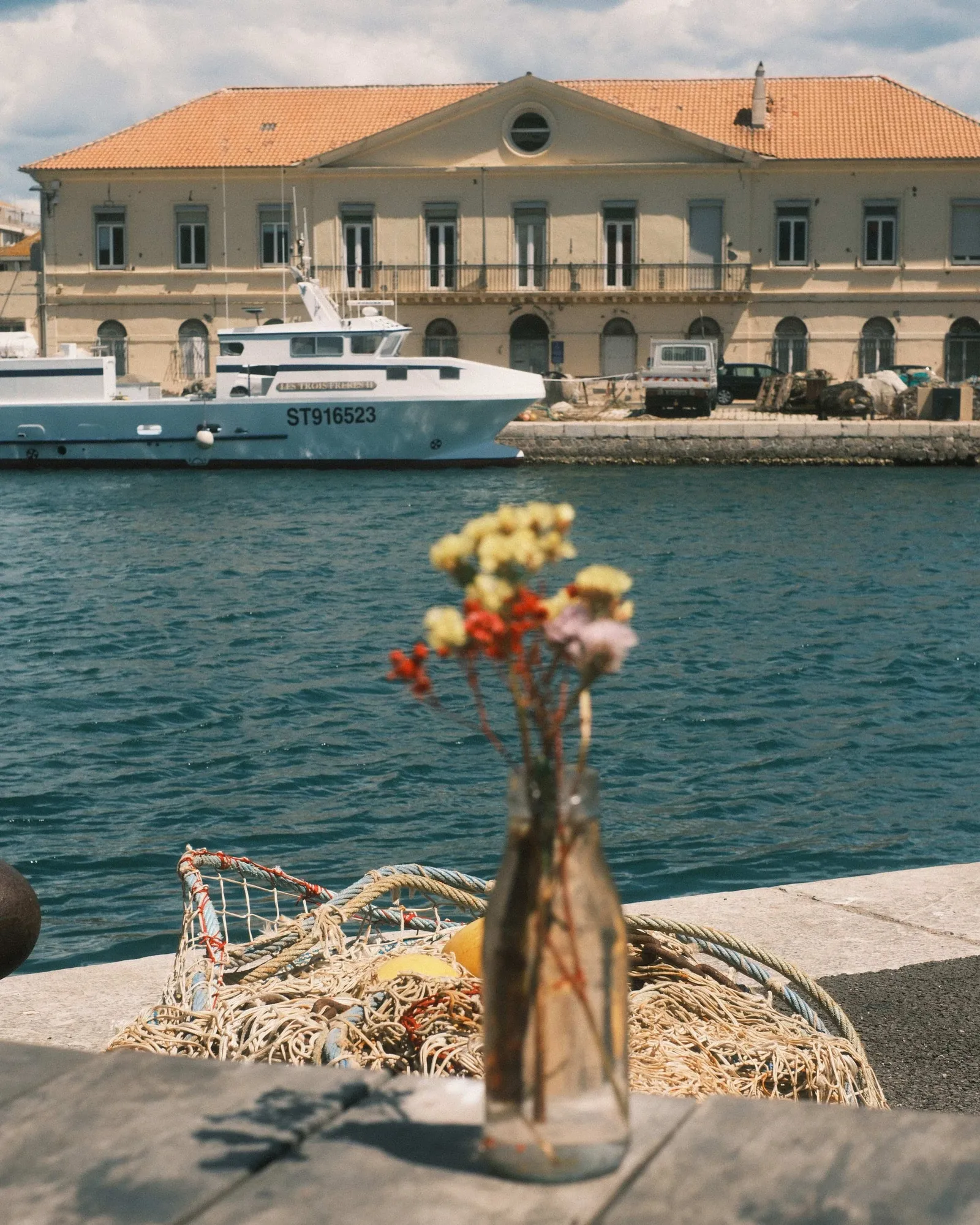 Wild flowers in a glass bottle on a quay in Sete, fishing nets and a trawler behind, shot with the Last Summer Roll recipe