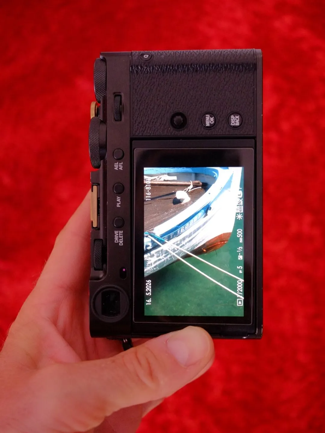 Back of the Fujifilm X-E5 held in one hand, rear screen showing a moored fishing boat frame shot in Sete