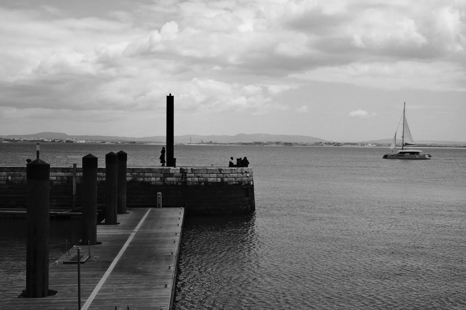 Black and white view of the Tagus from a wooden jetty, figures on the stone pier and a catamaran under sail