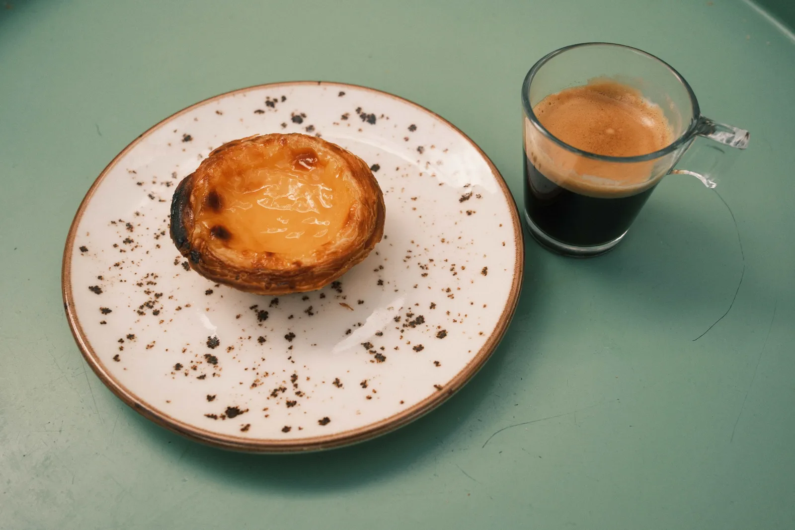 Pastel de nata dusted with cinnamon on a speckled plate beside an espresso on a green table