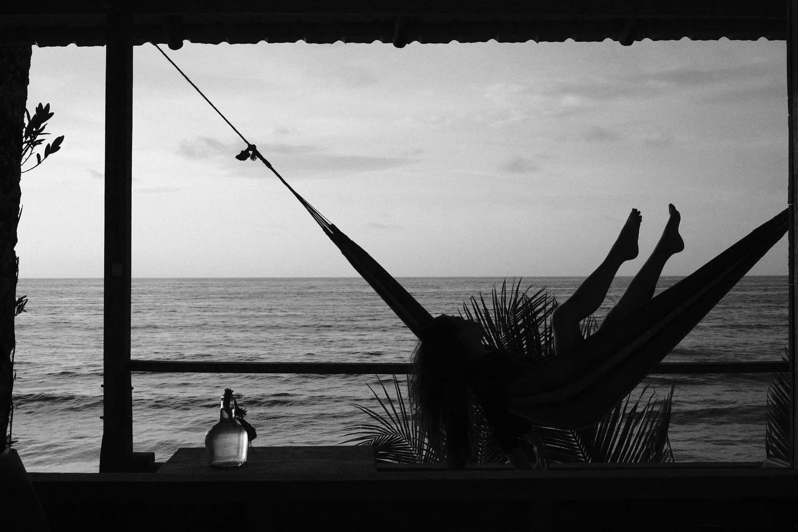Silhouette of a woman lying in a hammock facing the sea from a shaded terrace, black and white