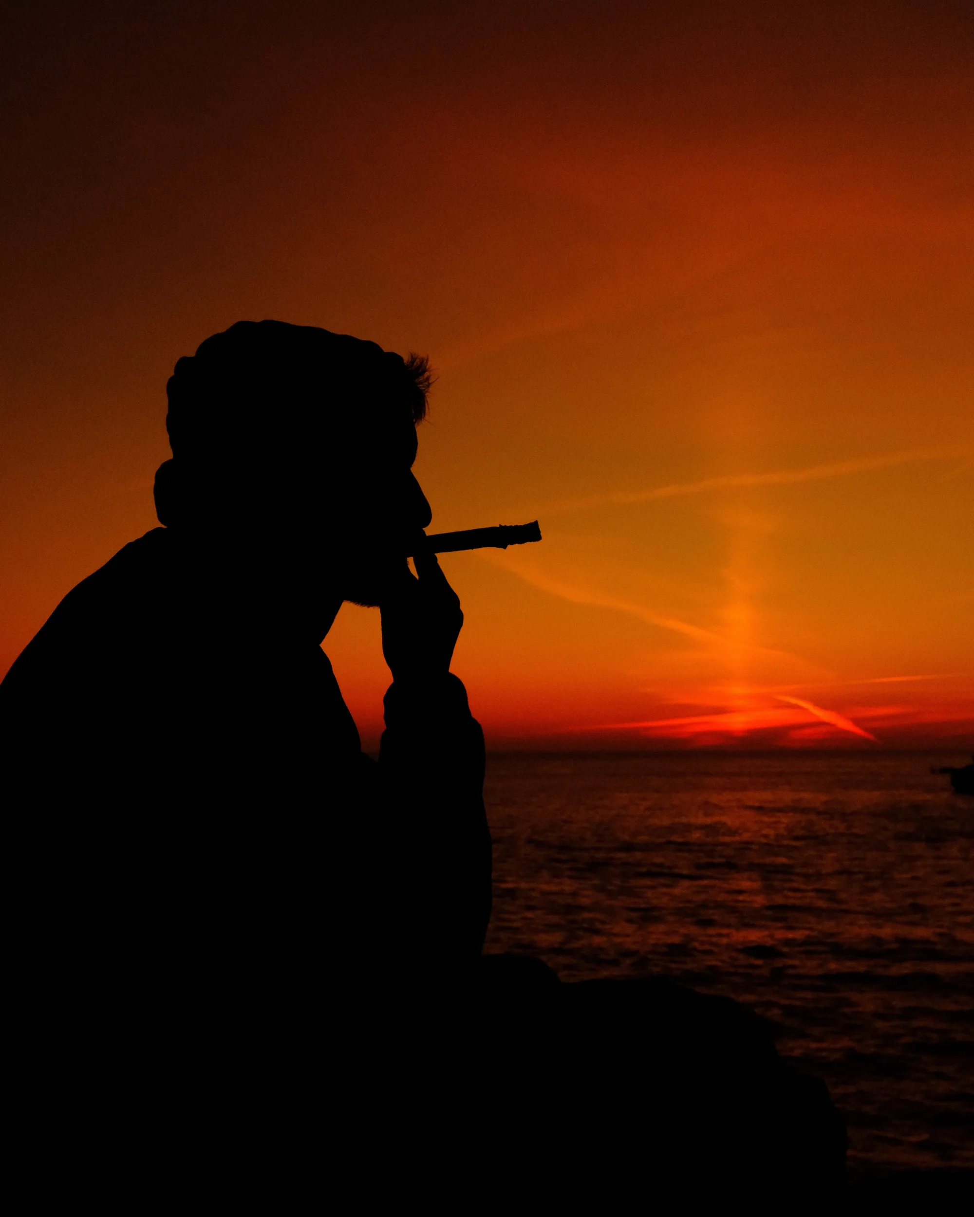 Close-up silhouette of a man smoking against a deep orange sunset over the water